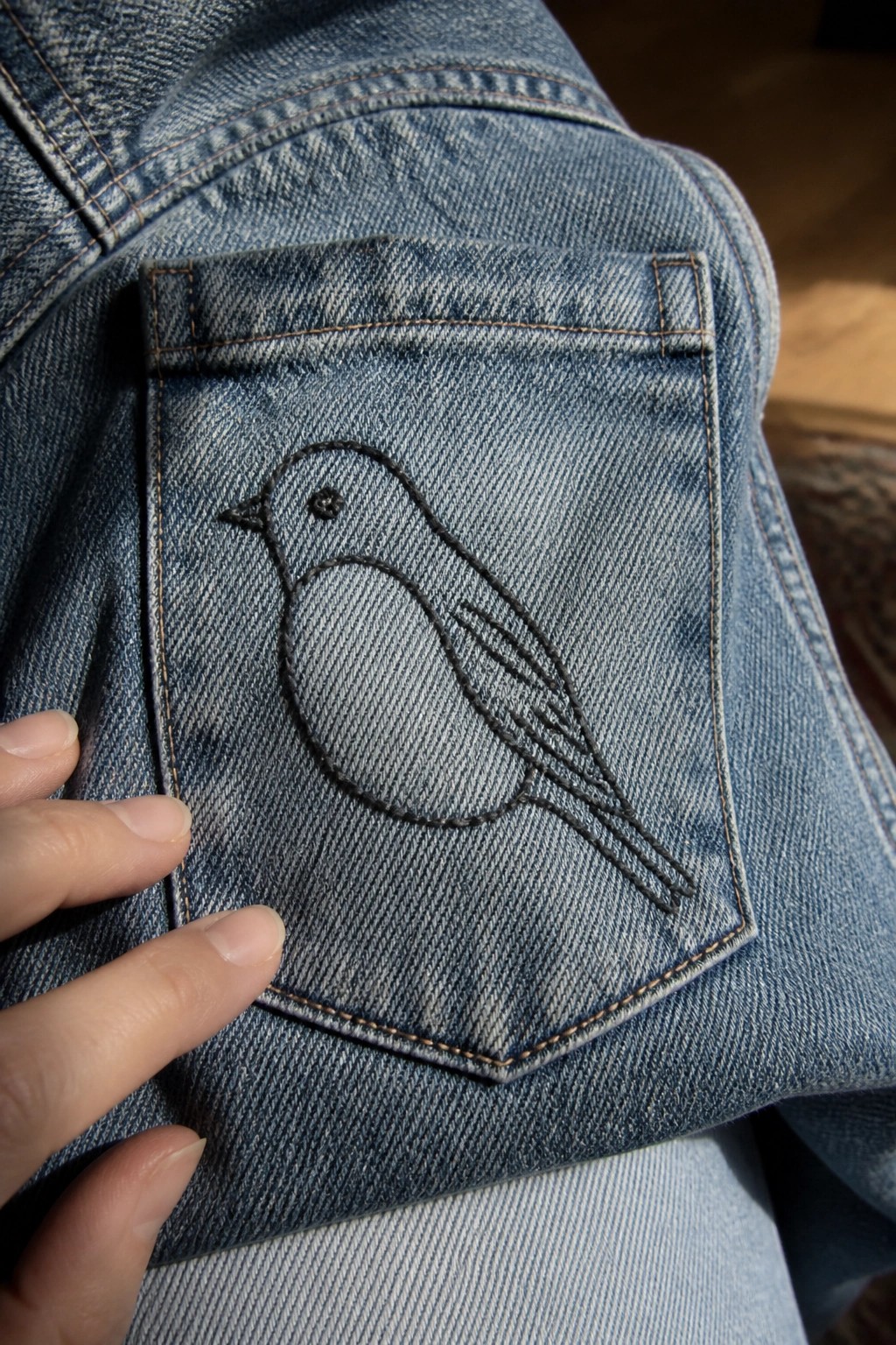 Blue denim pocket with black embroidered bird, hand touching fabric.