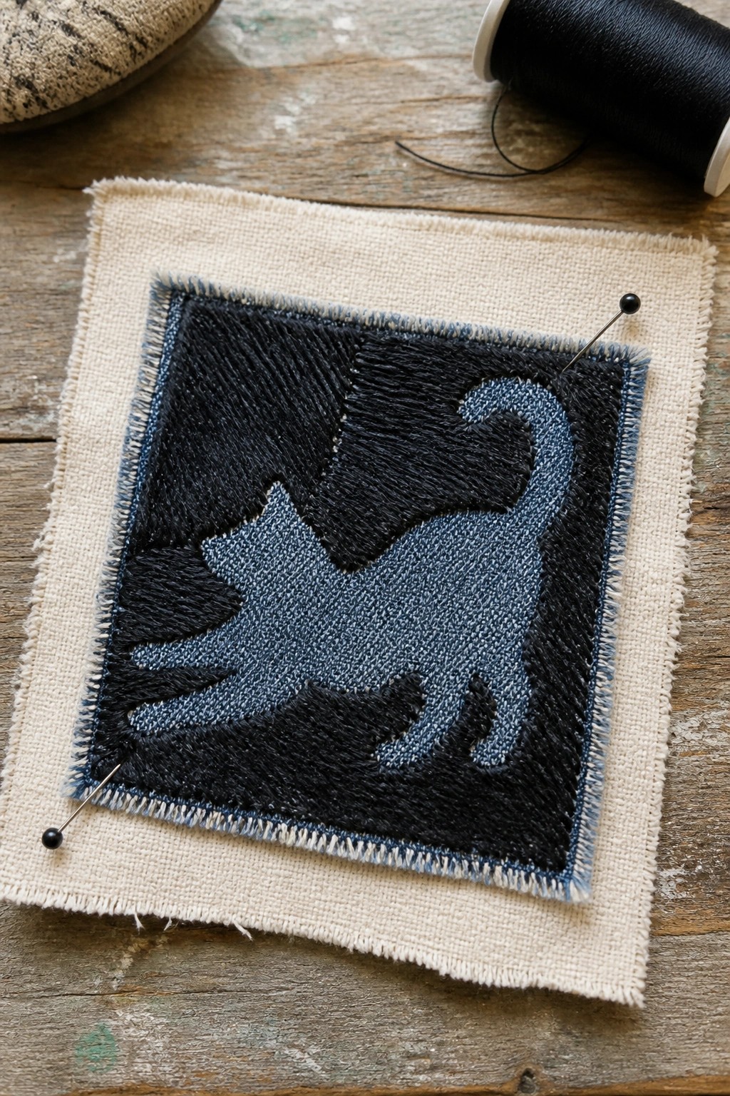 Denim cat appliqué in blue and black pinned to beige fabric on wood.