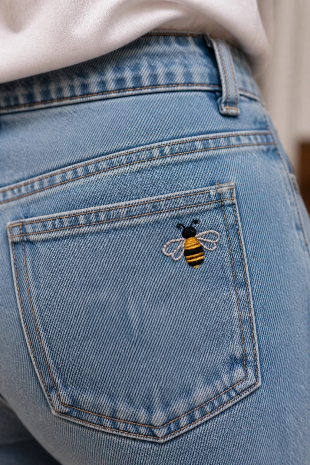 Close-up of light blue denim jeans back pocket with a small embroidered bee in black, yellow, and white threads.