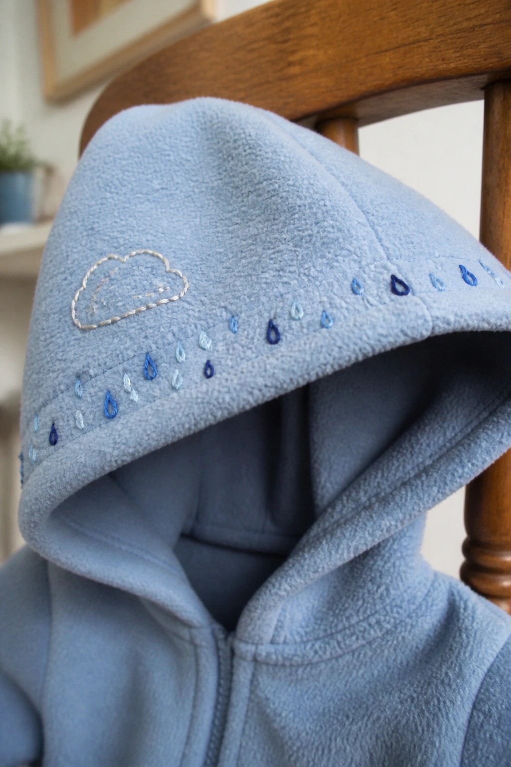 Light blue fleece hoodie with embroidered cloud and blue raindrops on wooden chair