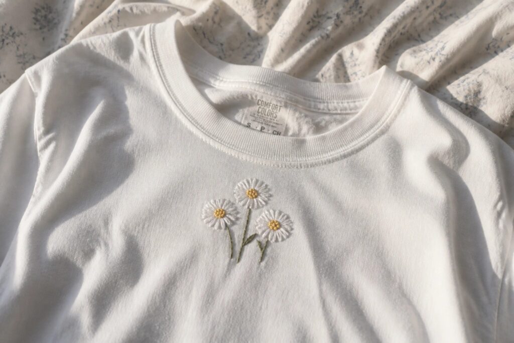 19 Easy Embroidery on Clothes Projects for Beginners White t-shirt with three embroidered daisies, white petals, yellow centers, green stems.