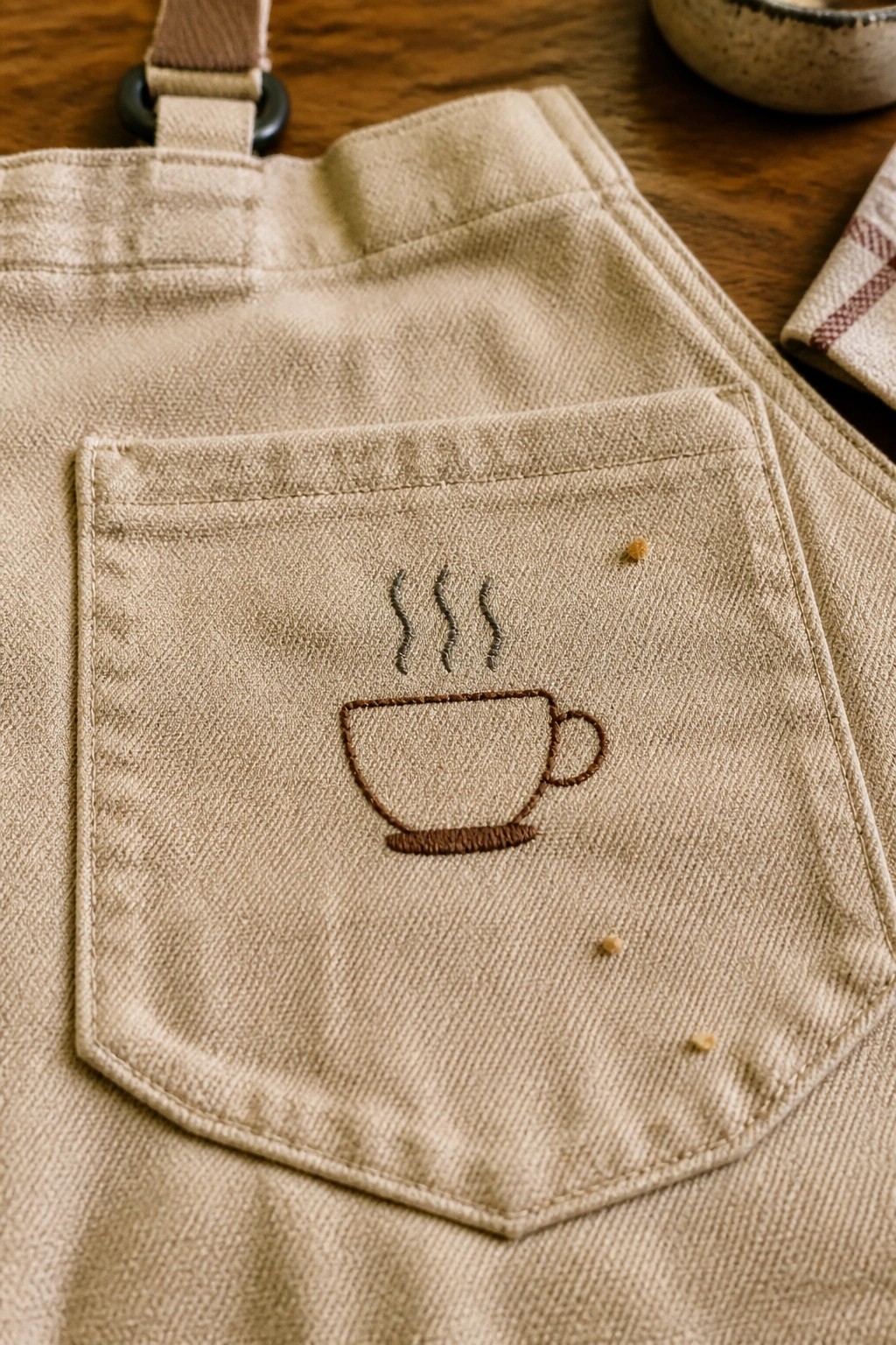Beige apron pocket embroidered with steaming brown coffee cup on wooden surface