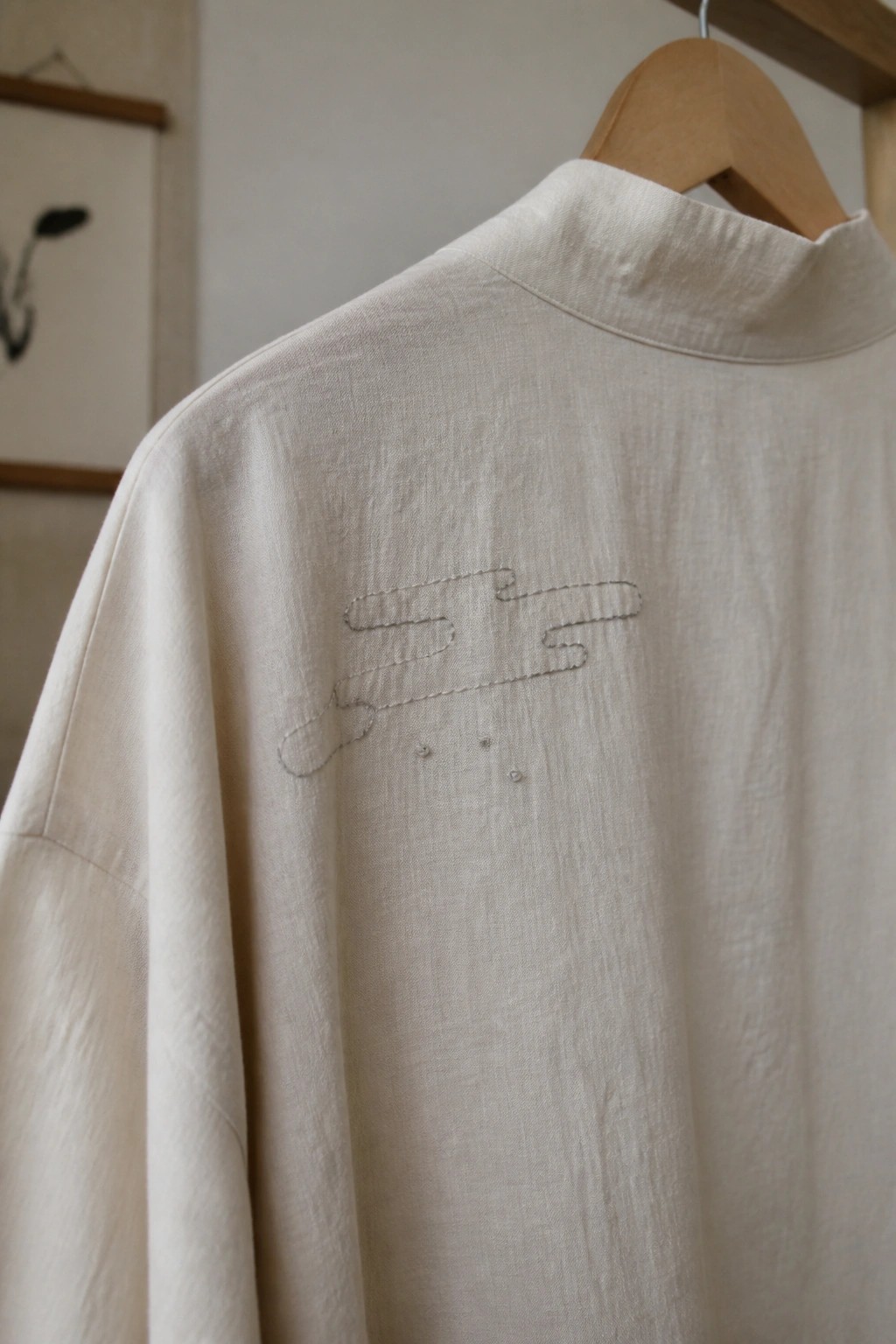Beige high-collared shirt with abstract embroidery on wooden hanger in room
