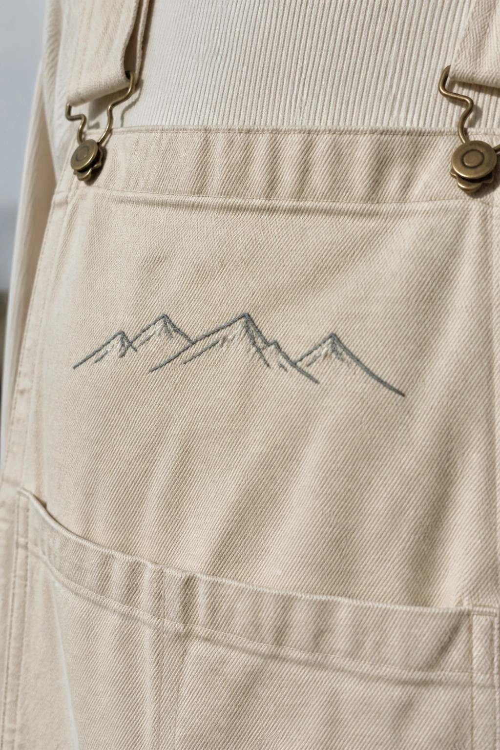 Beige overalls with gray embroidered mountain range above front pocket and brass clasps.