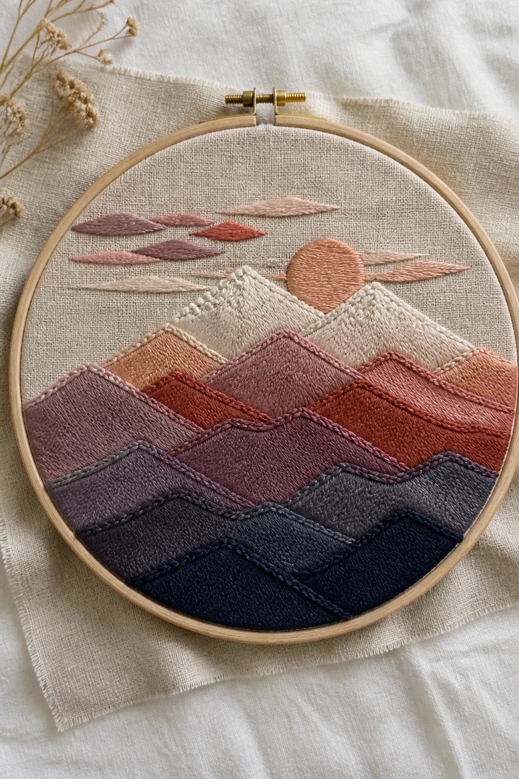 Circular embroidery hoop with layered pastel mountains, sun, and clouds on beige fabric.