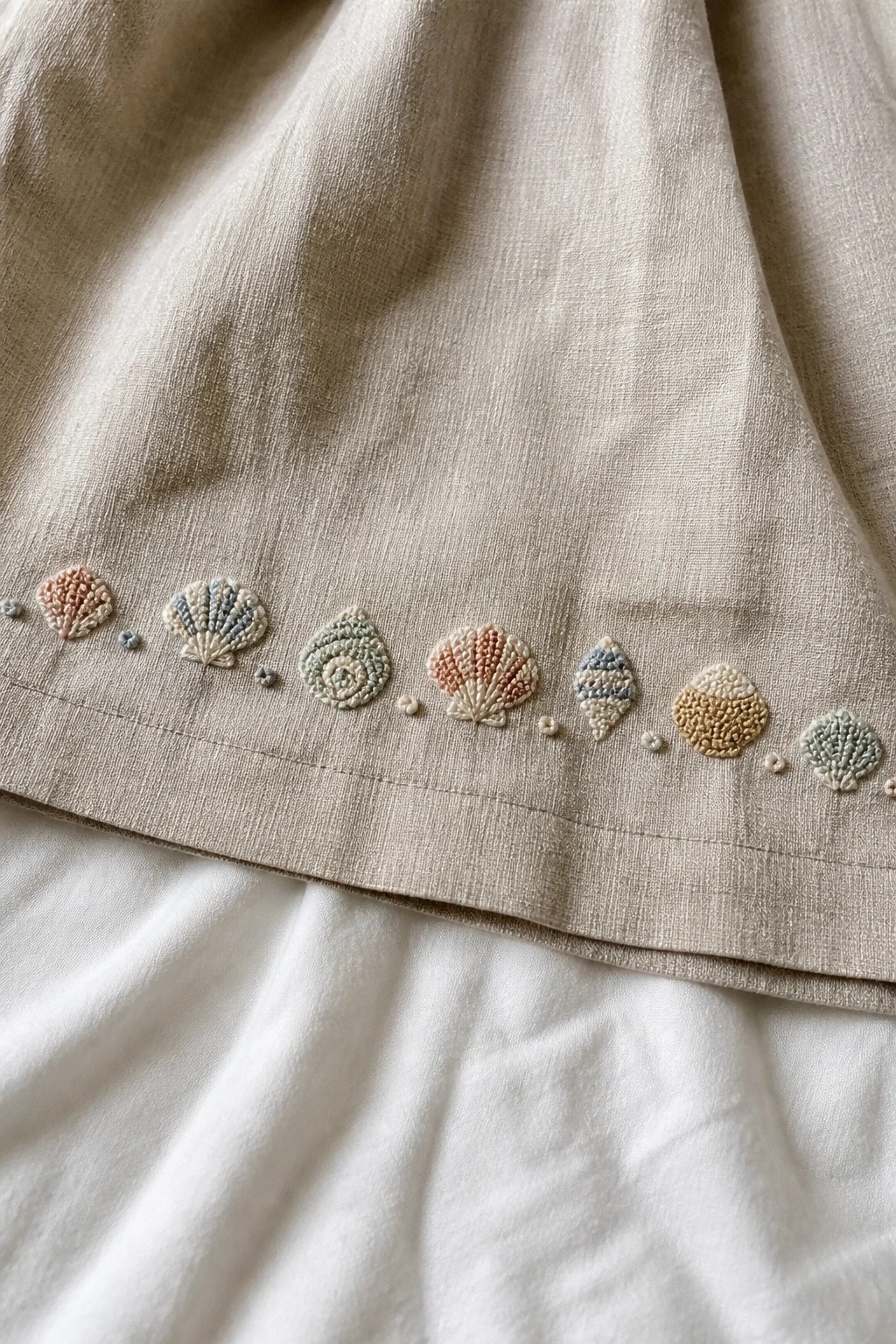 Beige linen garment hem embroidered with pastel seashells and dotted accents.