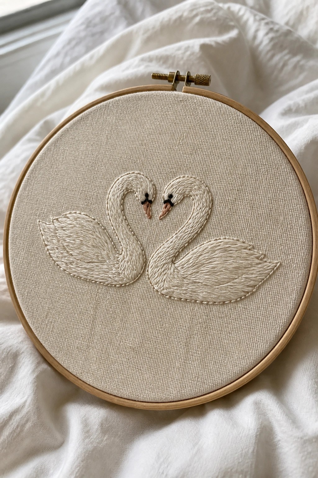 Two white swans with necks forming a heart, embroidered in a wooden hoop.