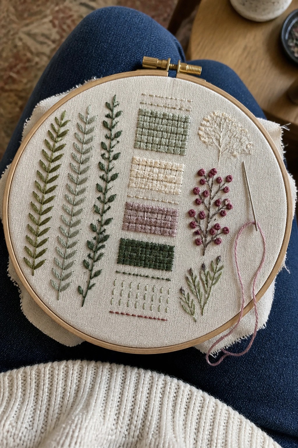 Wooden embroidery hoop with green leaves, purple berries, and stitches rests on blue jeans.