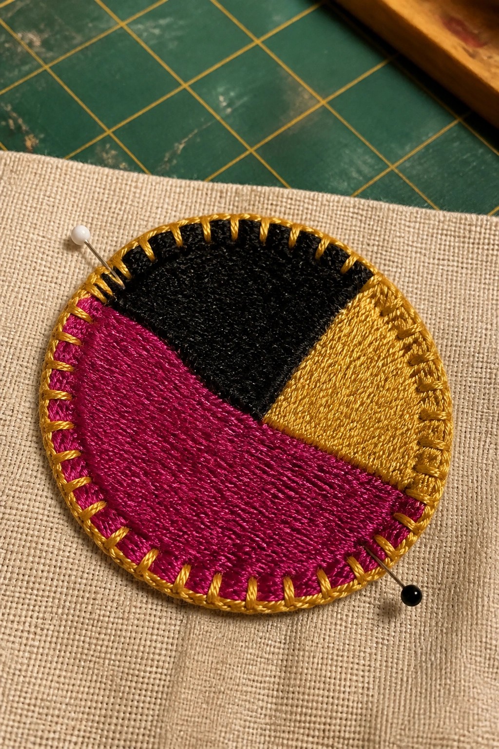Circular embroidered patch in magenta, black, and gold with gold border, pinned to fabric.