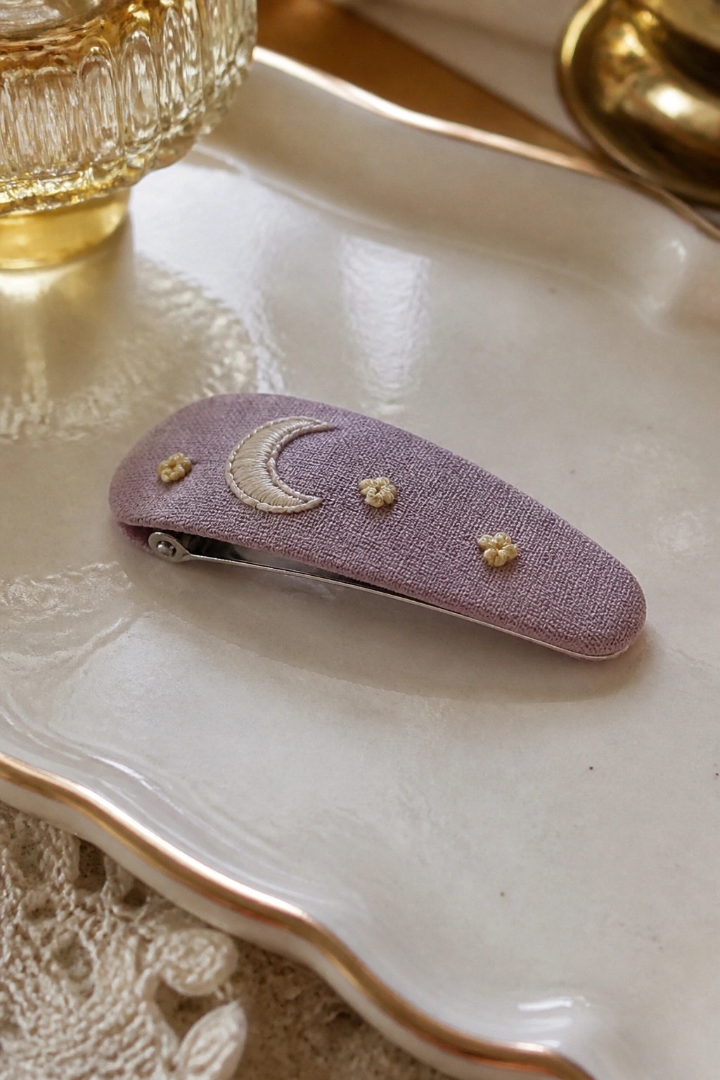 A lavender fabric hair clip with embroidered cream crescent moon and small floral motifs on a white tray.