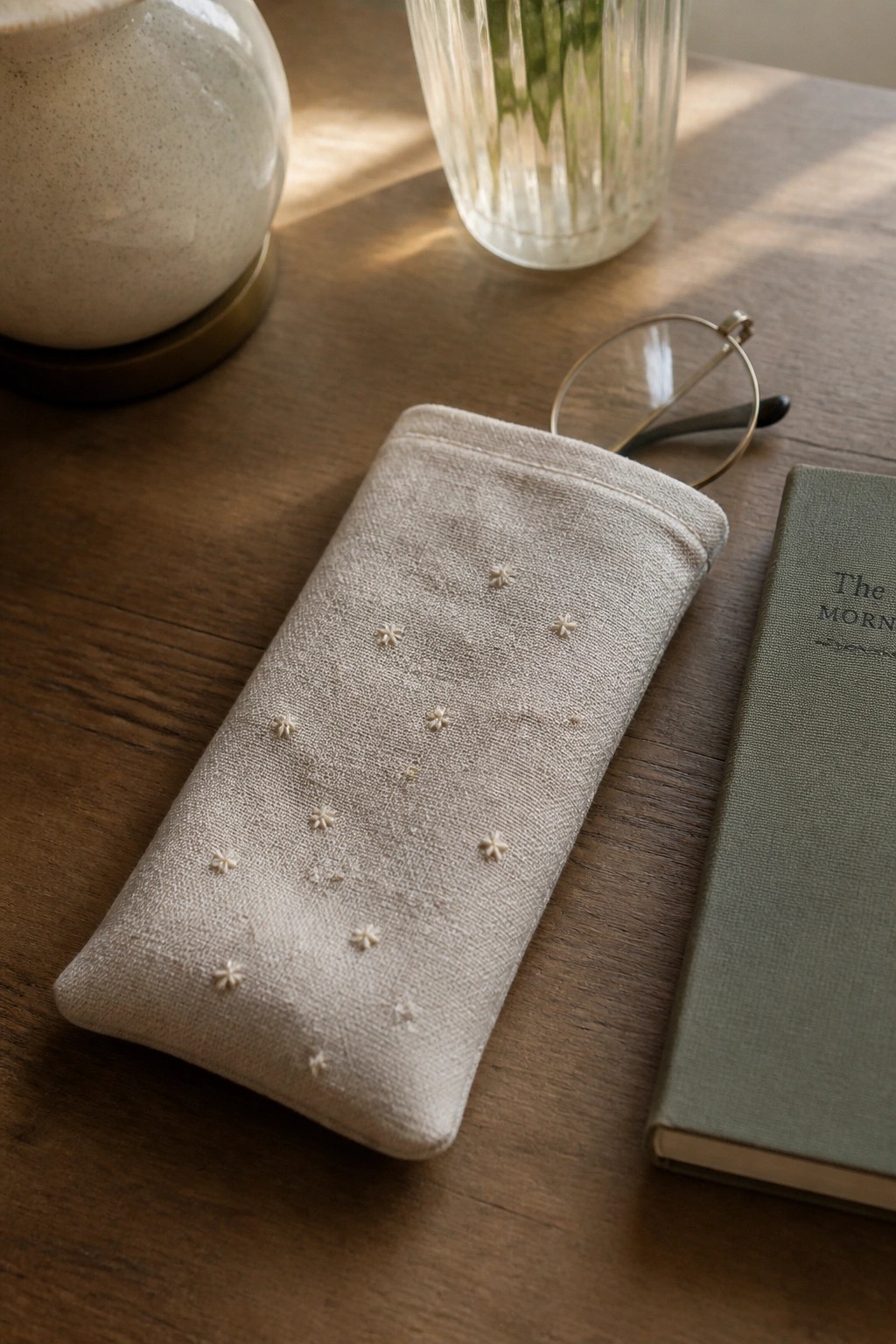 Embroidered beige linen pouch on wooden table beside eyeglasses and green book