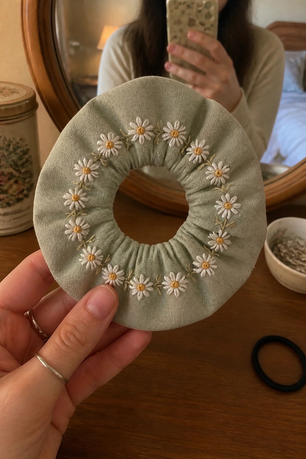 Hand holding sage green scrunchie embroidered with white daisies and yellow centers.