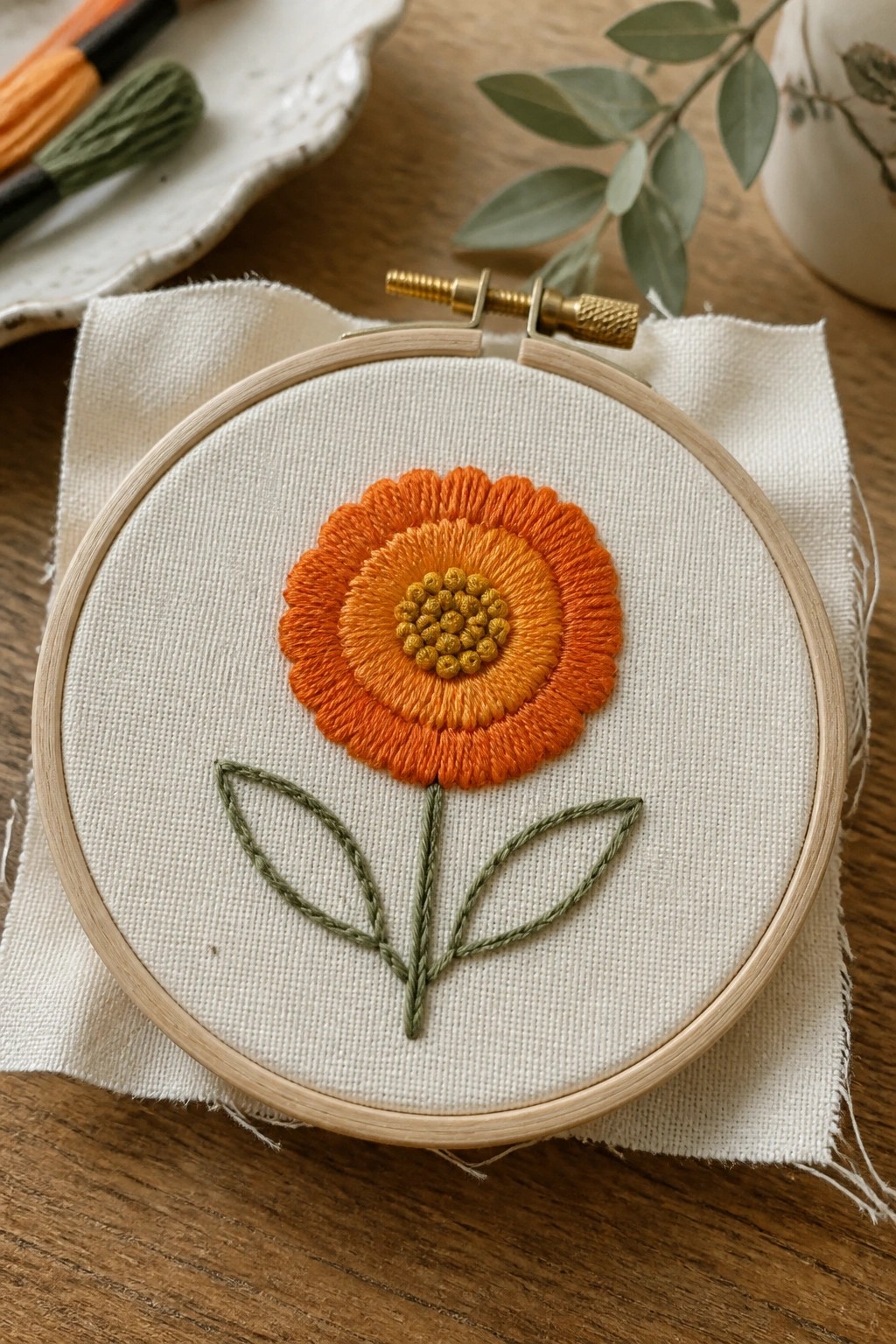 Orange and yellow flower embroidered on white fabric in wooden hoop with green leaves