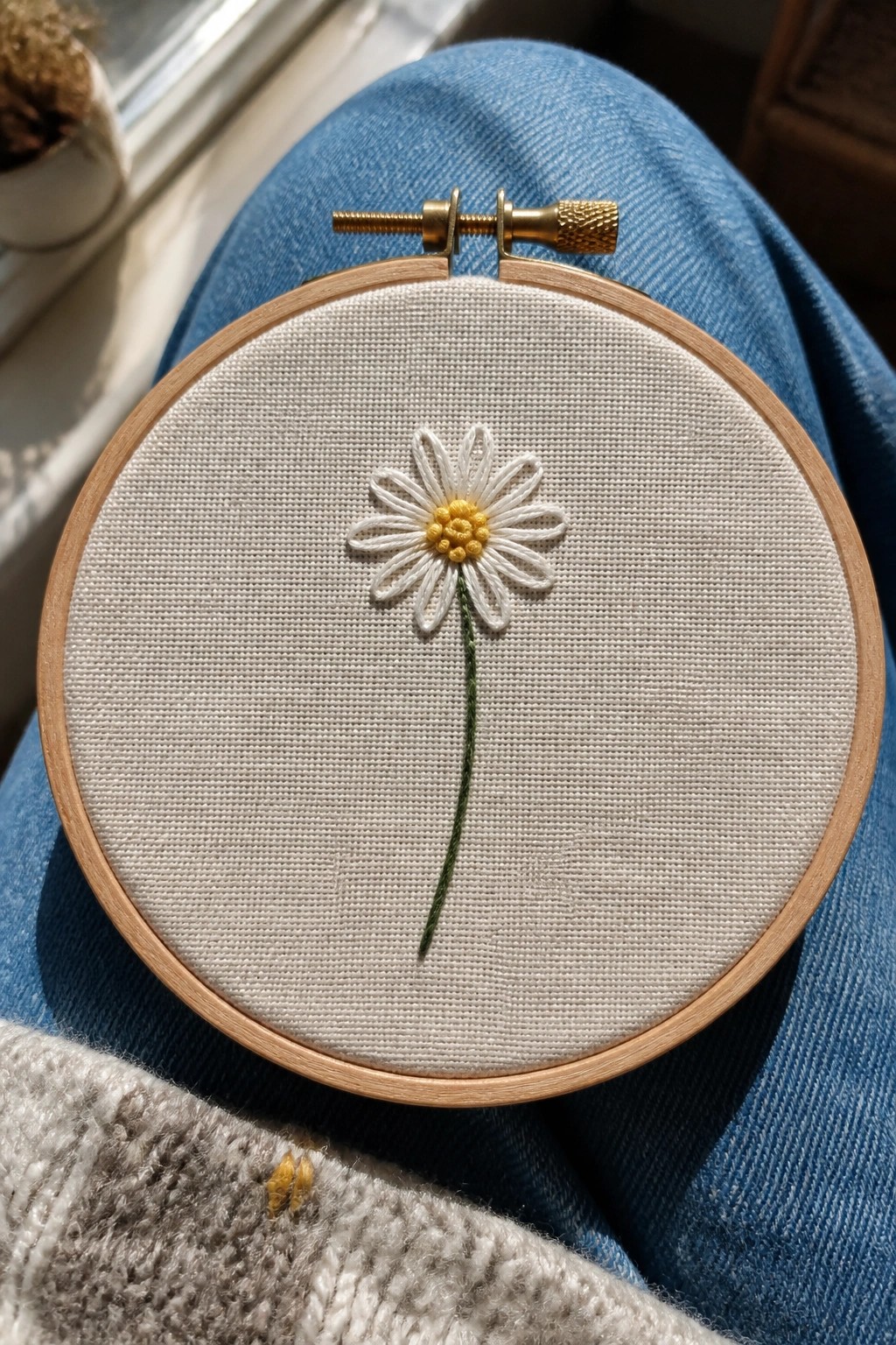 Wooden hoop with white daisy embroidery and green stem on blue denim jeans