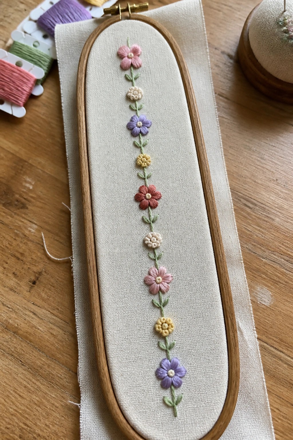 Wooden hoop with vertical pastel floral embroidery on beige fabric amid thread spools