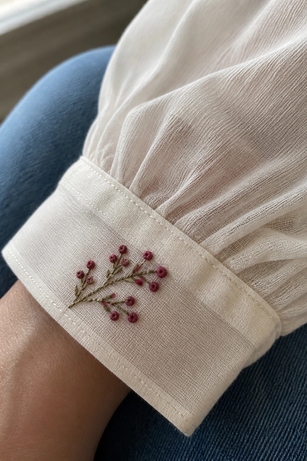 White cuff embroidered with pink berries on green stems, over blue jeans.