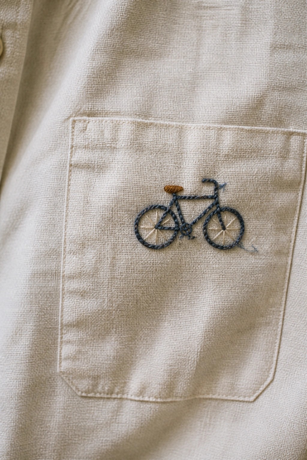 Small embroidered bicycle centered on the pocket of a beige shirt.