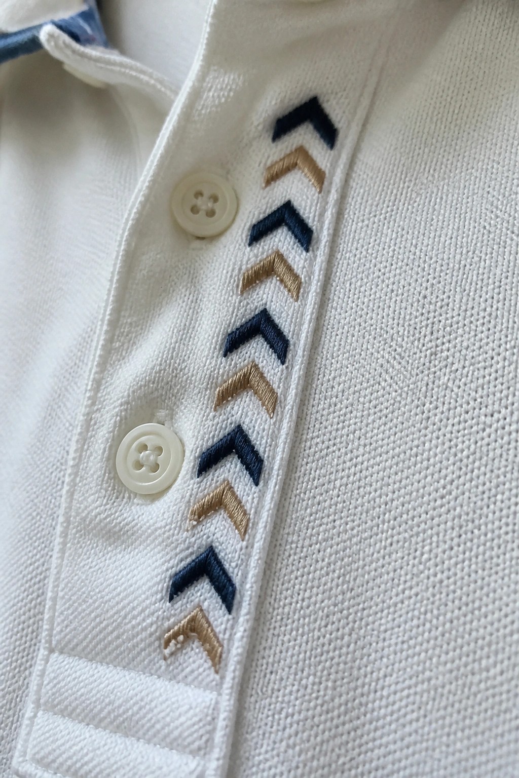 White polo shirt placket with navy and gold chevron embroidery.