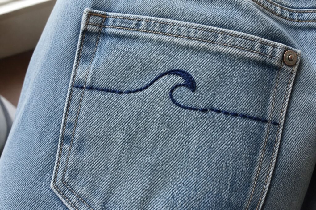 19 Simple Embroidery Ideas for Clothes Using Basic Stitch Techniques Blue denim jeans pocket with dark blue wave embroidery and orange stitching.