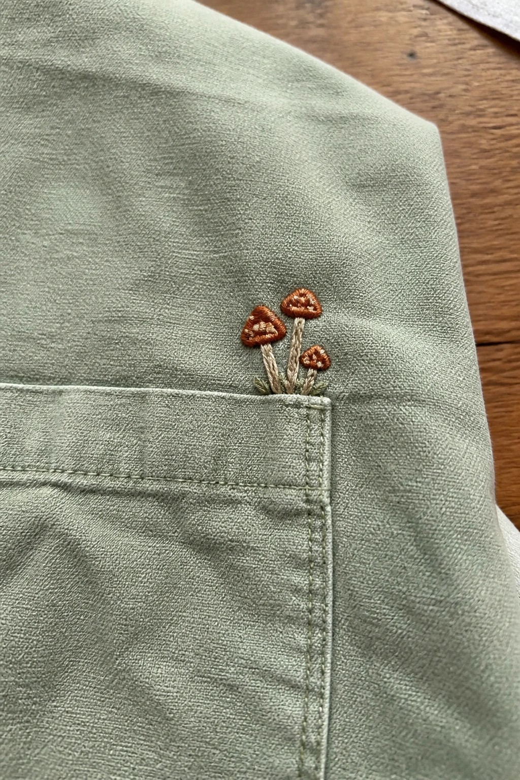 Sage green fabric pocket embroidered with three small brown mushrooms