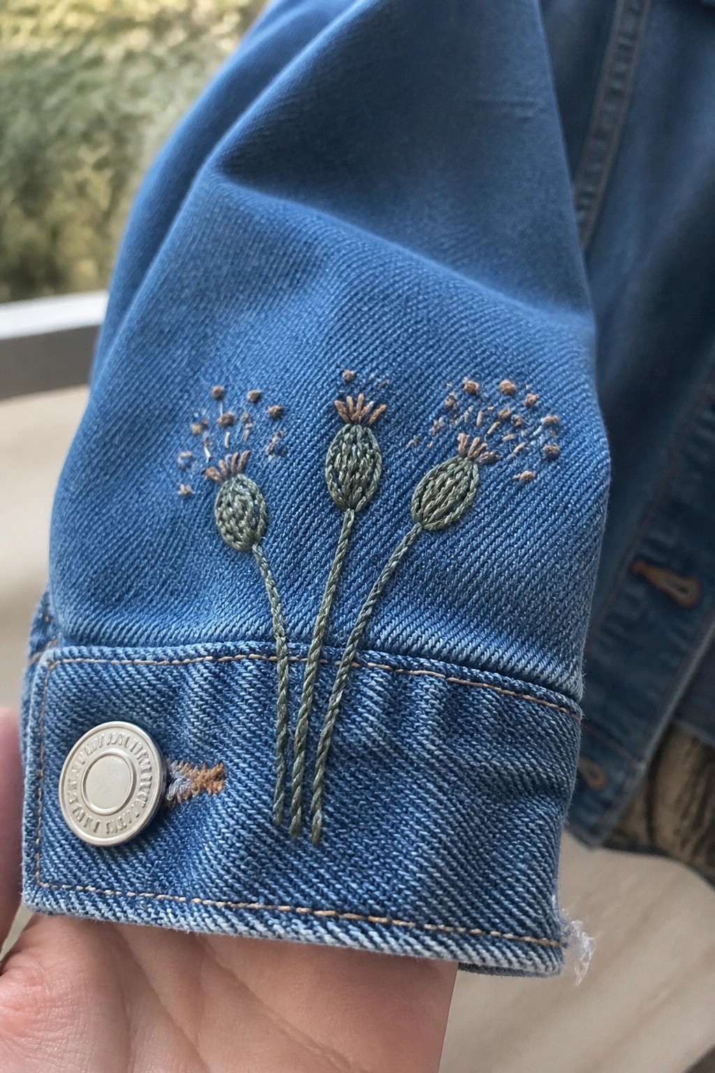 Blue denim jacket cuff with green and brown embroidered seed heads beside a button.