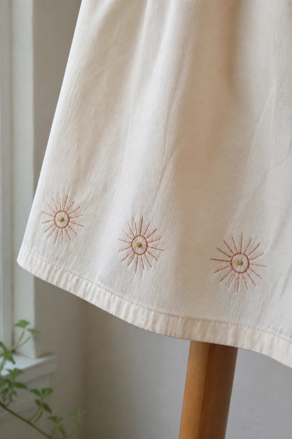 Off-white skirt hem with three orange embroidered sun motifs on wooden stand