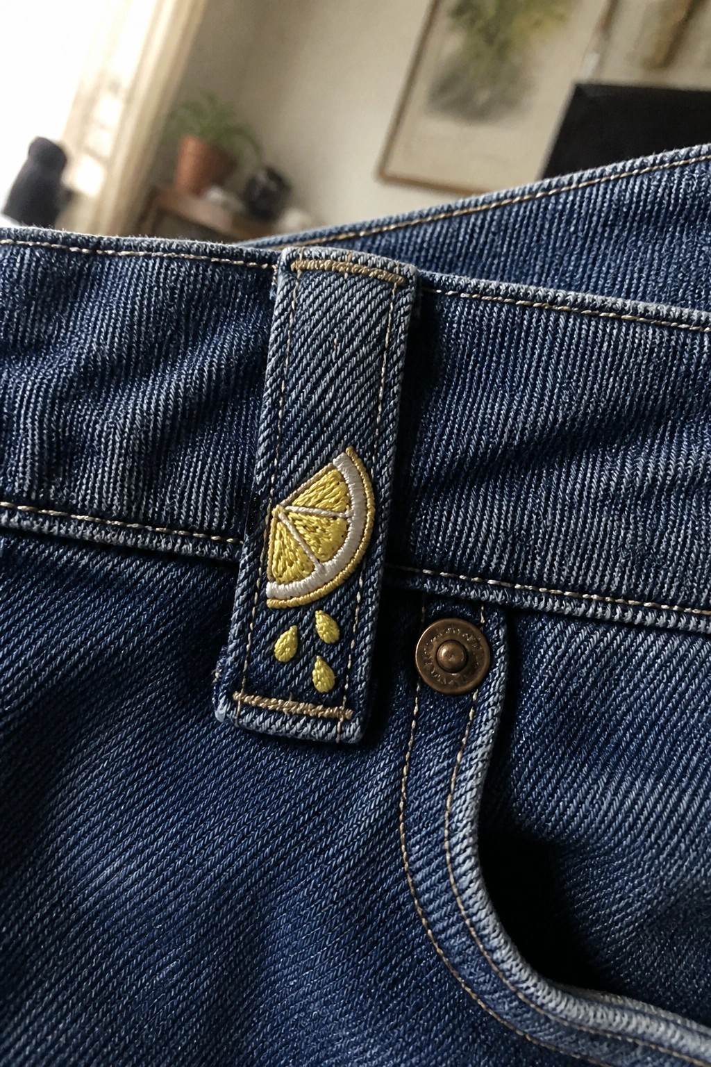 Blue denim jeans with embroidered yellow lemon on belt loop beside brass button