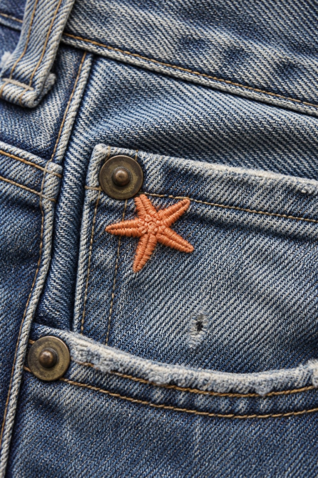 Blue denim jeans pocket featuring orange embroidered starfish beside metal rivet button.