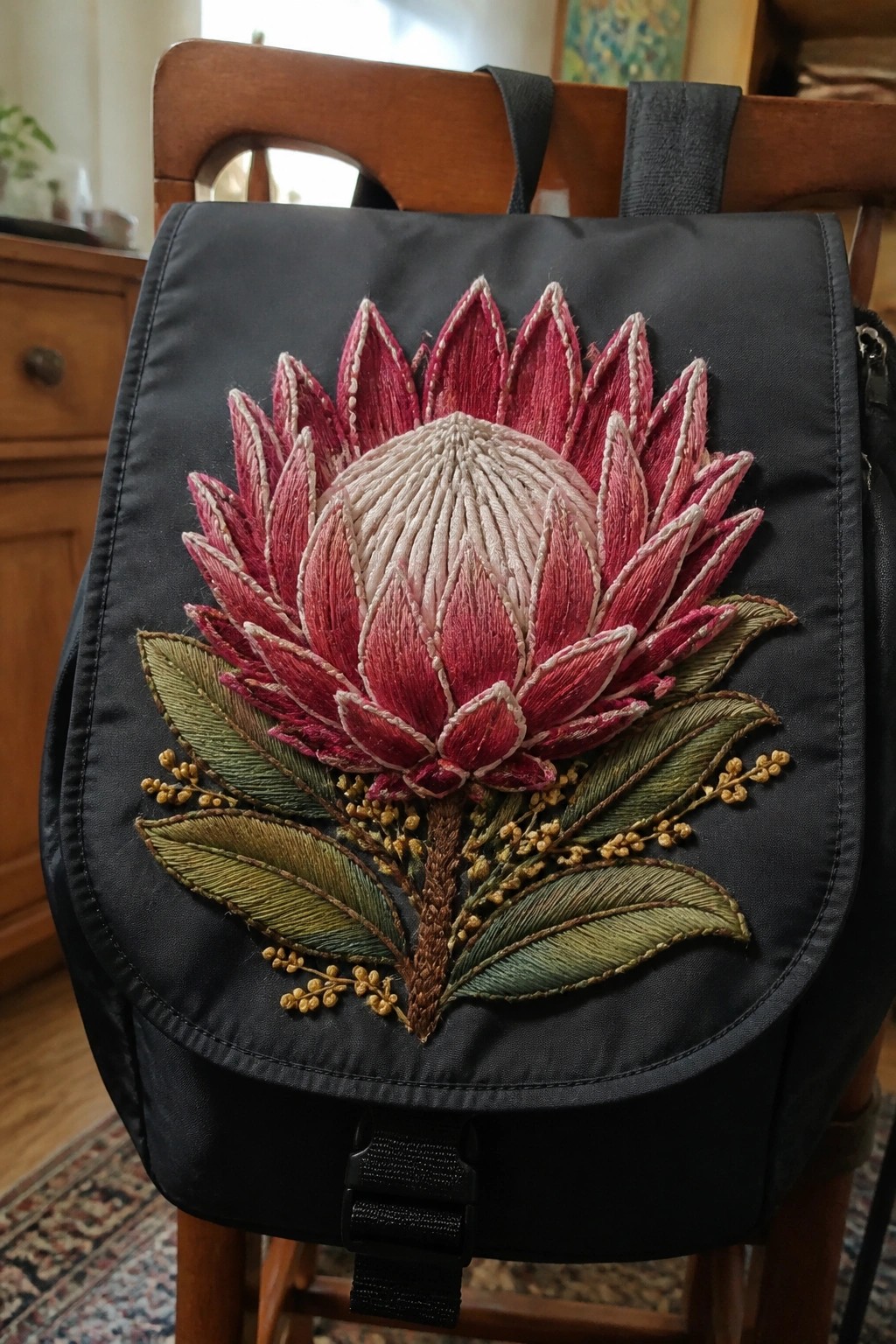 Black backpack with embroidered pink flower, green leaves, and yellow buds on wooden chair