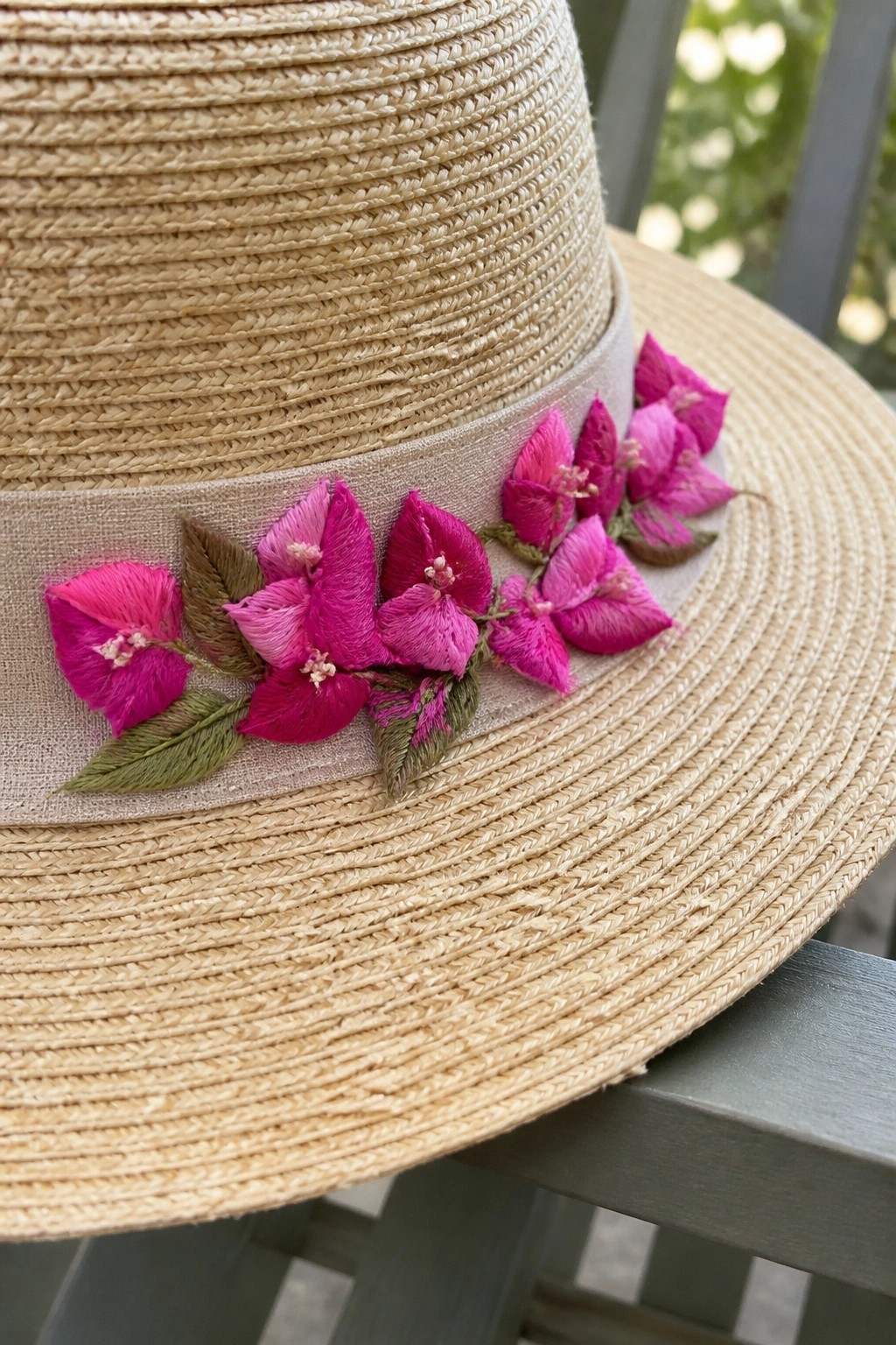 Close-up of a straw hat with a beige ribbon band embroidered with clusters of bright pink flowers and green leaves.