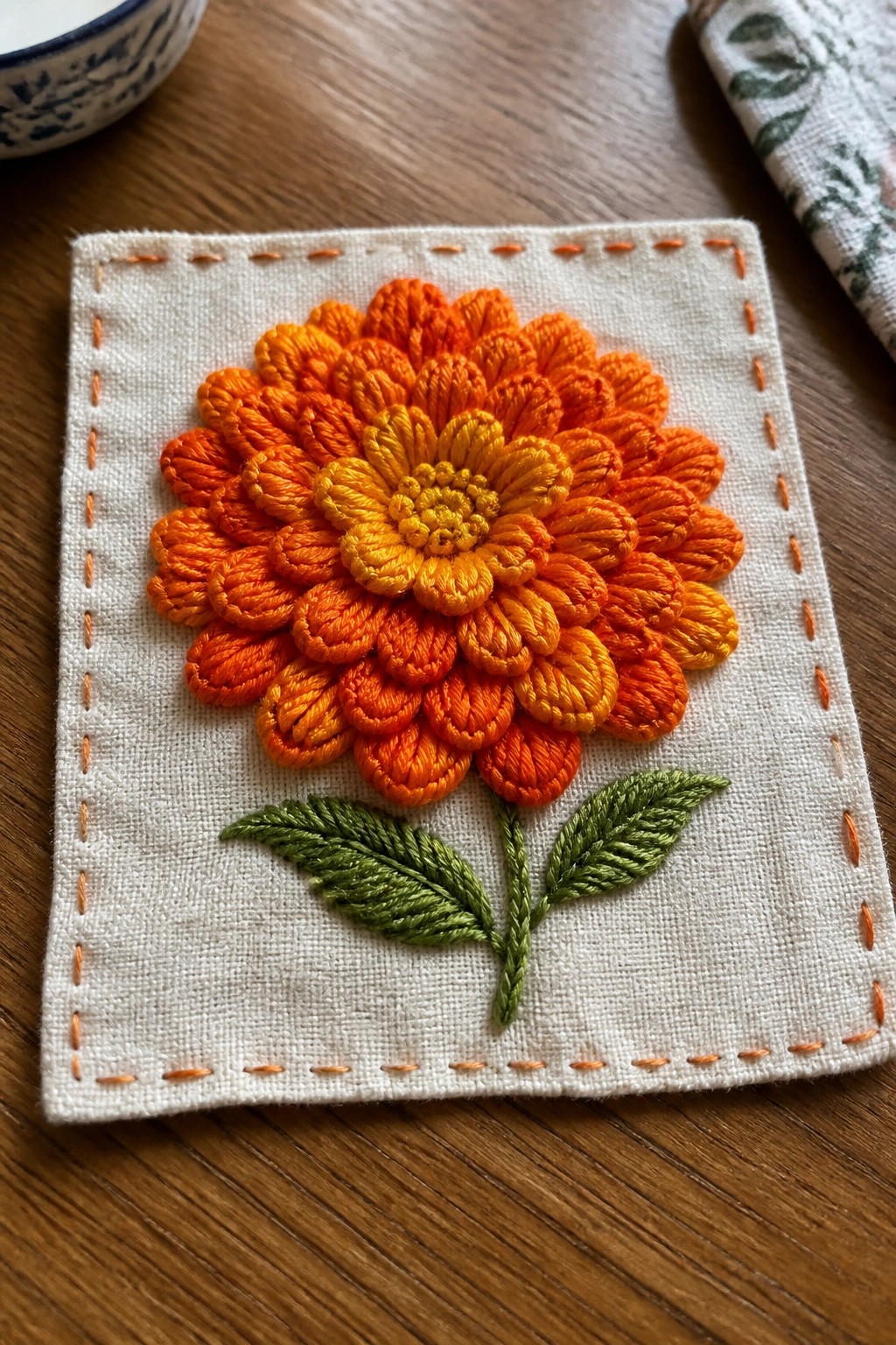 Embroidered orange-yellow flower with green leaves on white fabric with orange border