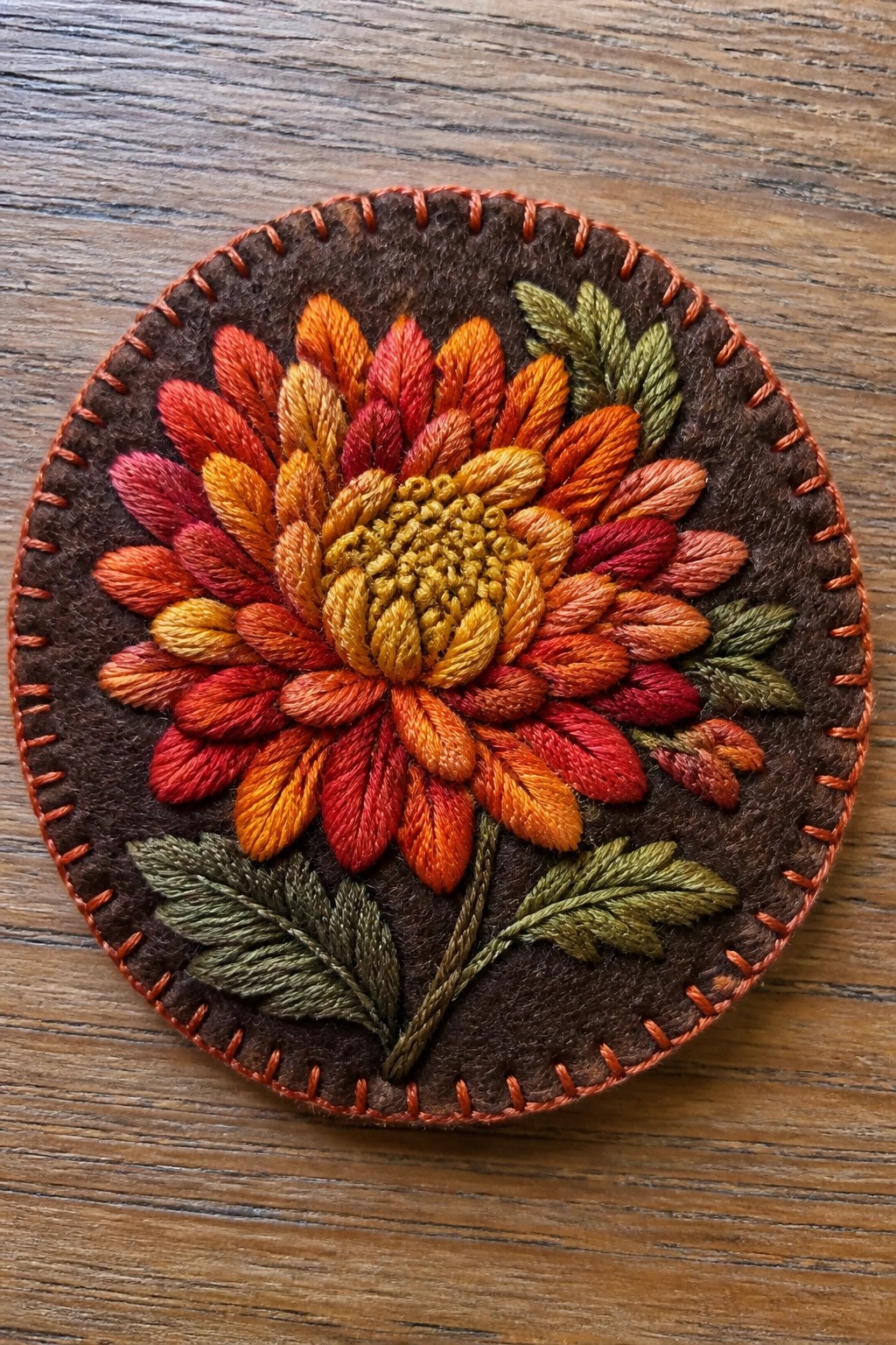 Round felt embroidery of vibrant red-orange flower with green leaves on wood.