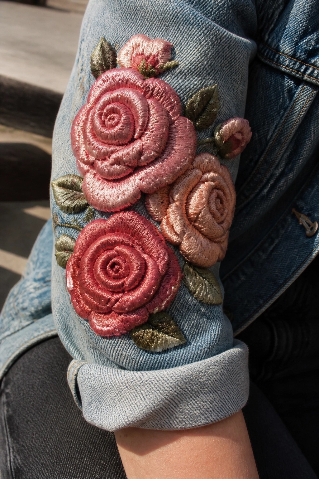 Denim jacket sleeve embroidered with large pink roses and rolled cuff.