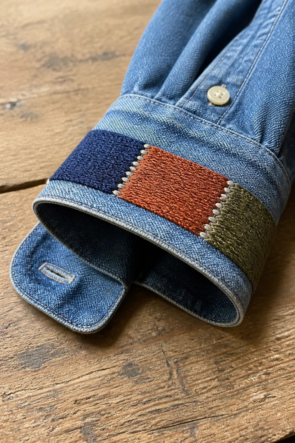 Denim cuff with navy, orange, and green fabric patches stitched in white on wood.