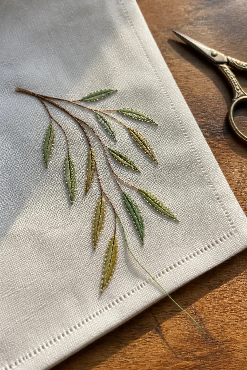Embroidered green and gold leaves on brown stems over white fabric beside scissors.