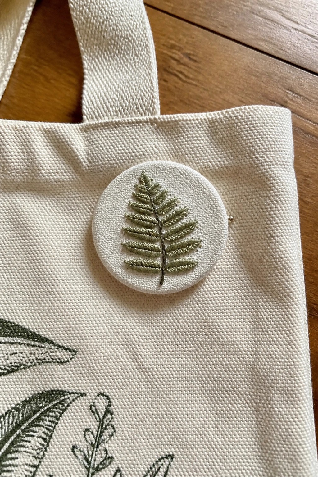 White canvas tote bag with circular green fern-embroidered brooch pinned on front.