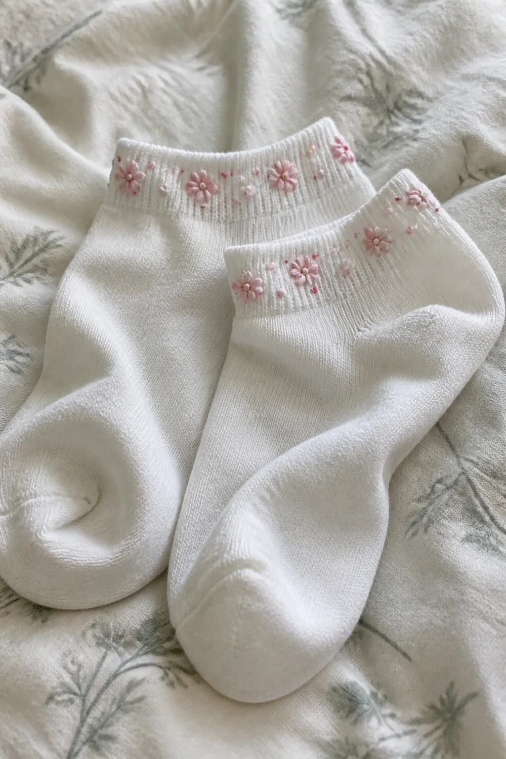 Two white ankle socks with pink floral embroidery on patterned green fabric.