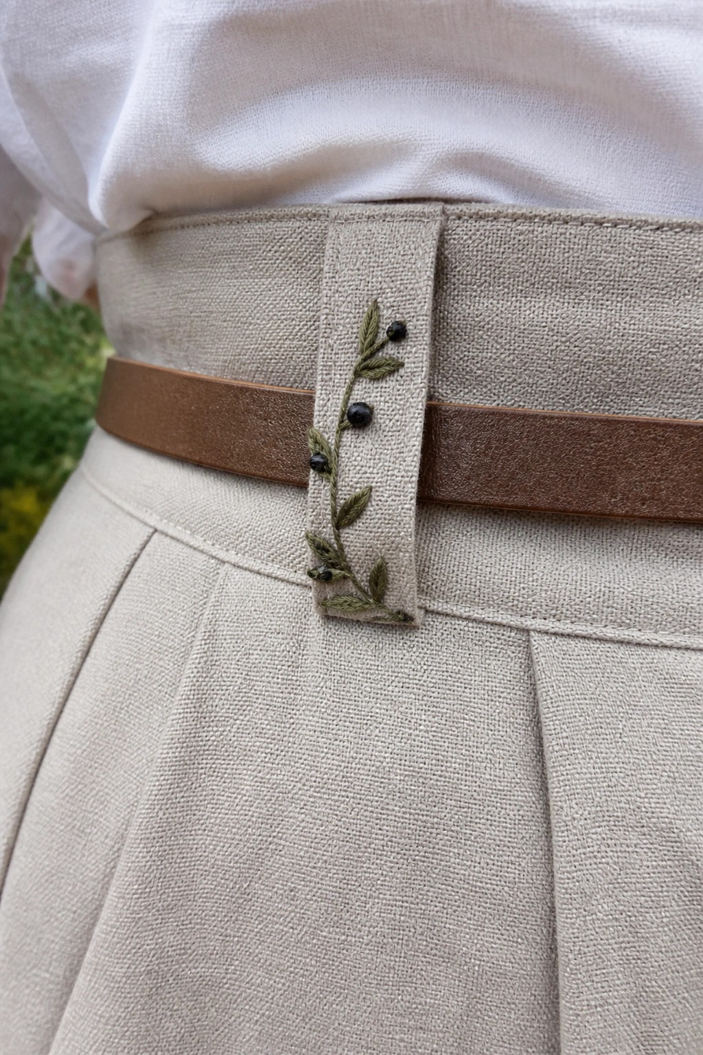 Beige pleated skirt with brown belt and embroidered vine on tab.