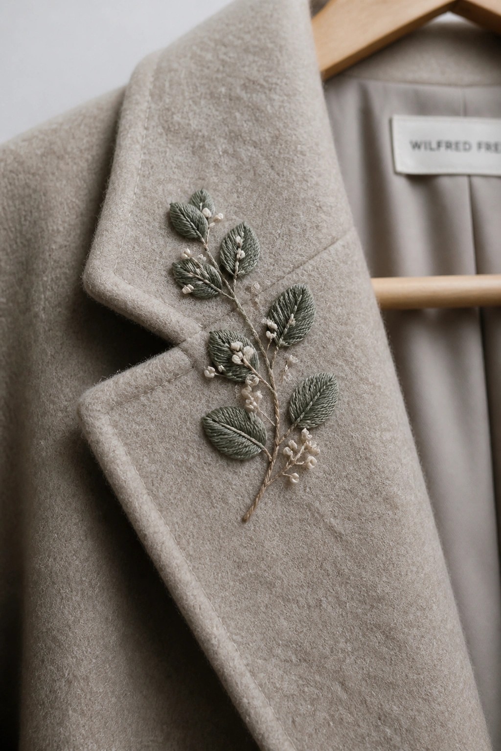Close-up of a beige wool coat lapel with green embroidered leaves and small berry clusters along the edge.