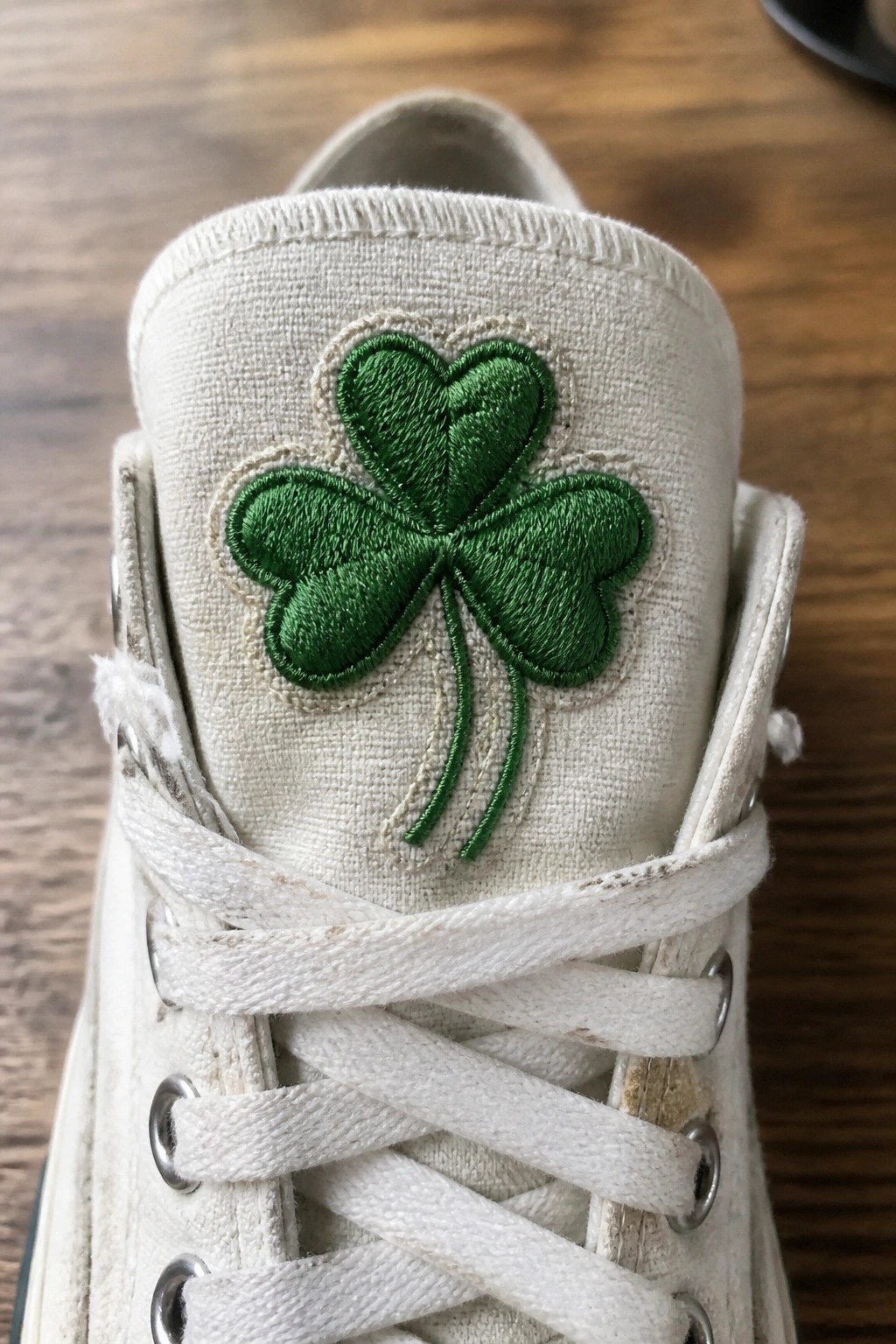 White canvas sneaker tongue with green embroidered three-leaf clover above white laces.