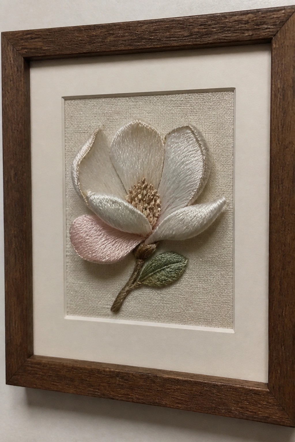 Embroidered white magnolia with pink petal, green leaf, in dark wooden frame