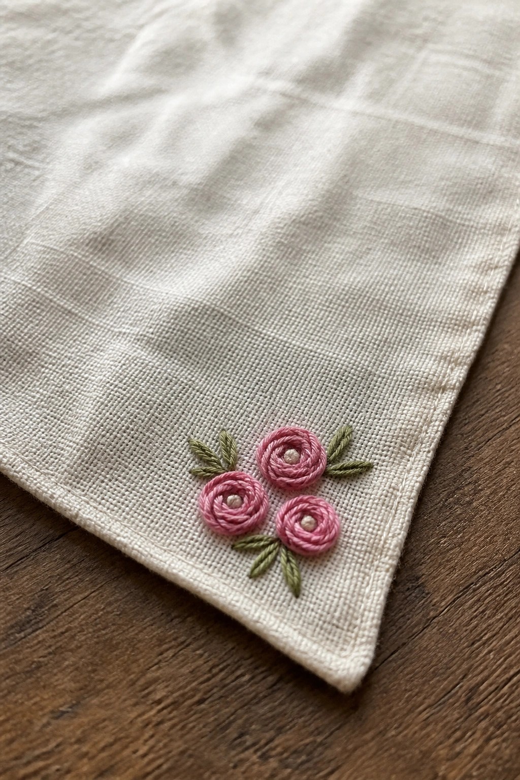 White fabric corner with three pink rose embroideries and green leaves on wood