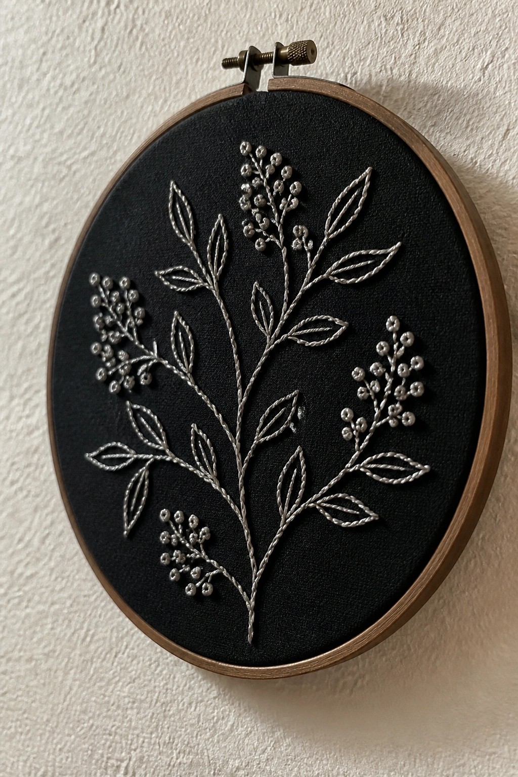 Silver-stitched botanical design with leaves and berries on black fabric in wooden hoop.