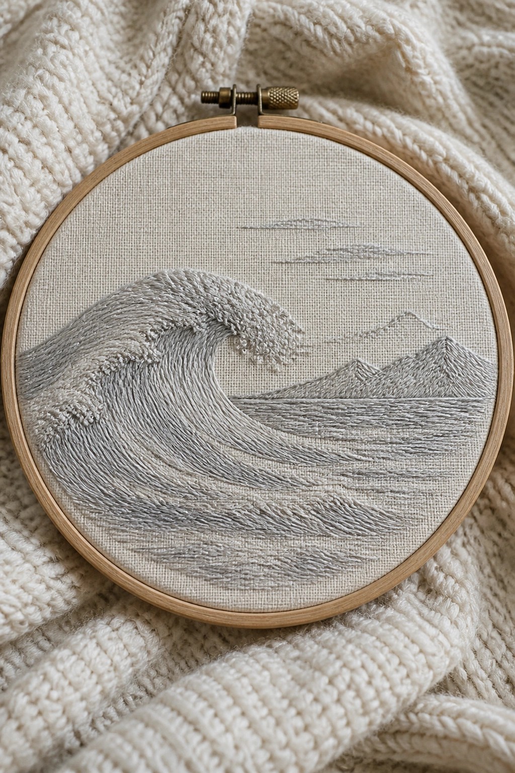 Wooden hoop with embroidered gray wave, mountains, and clouds on beige fabric over knit.