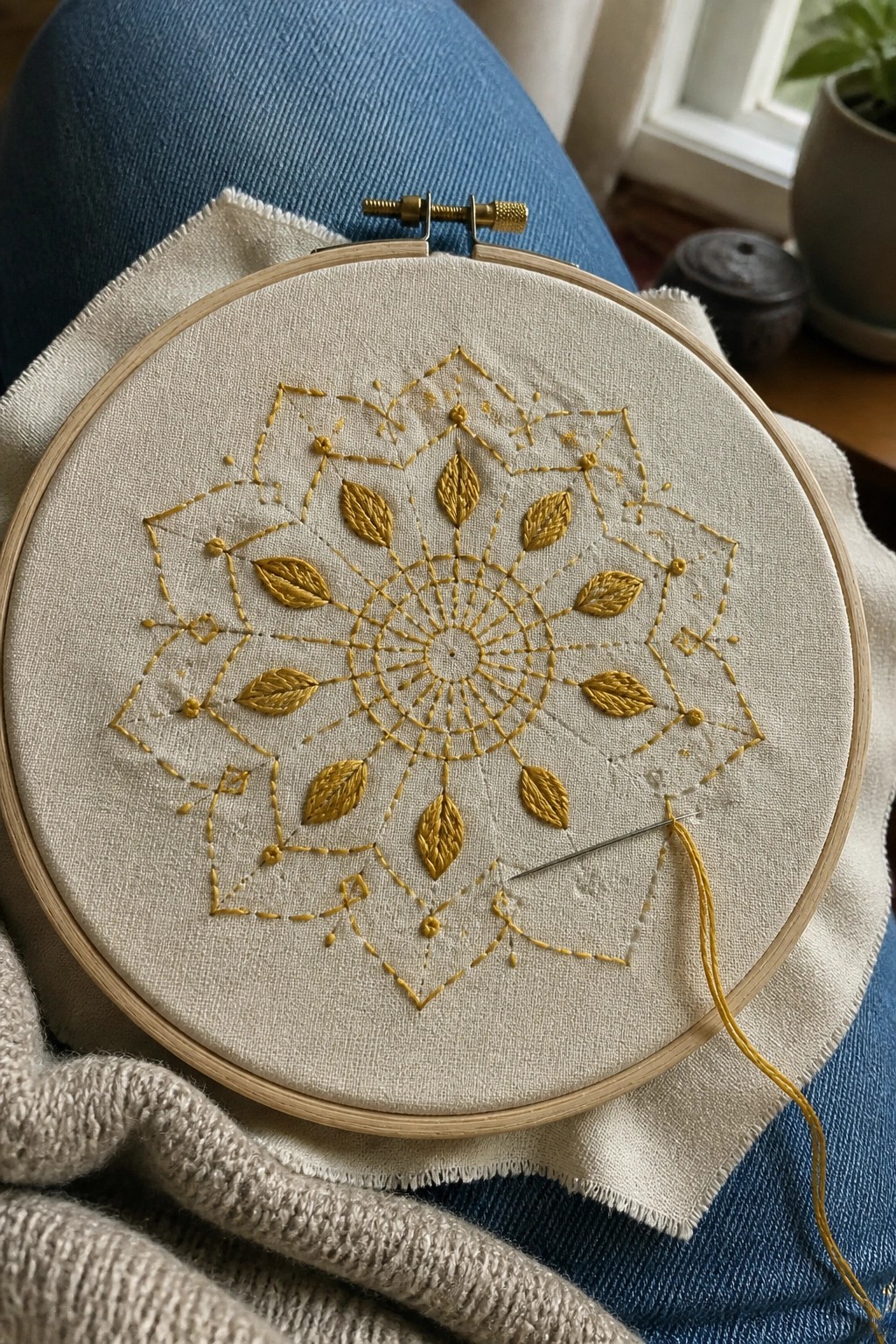 Wooden hoop with yellow mandala embroidery on white fabric, needle on blue jeans lap.