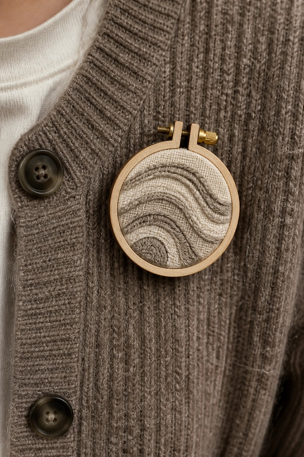 Knitted brown cardigan with round wooden hoop brooch displaying wavy beige embroidery.