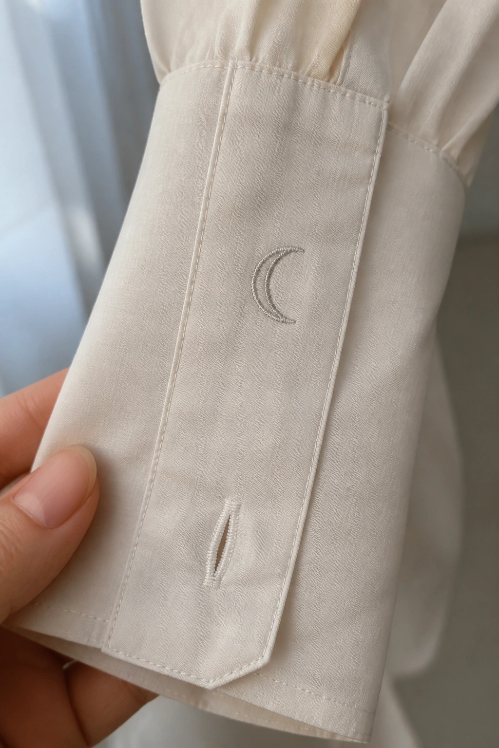 Hand holding off-white shirt cuff with silver crescent moon embroidery above buttonhole.