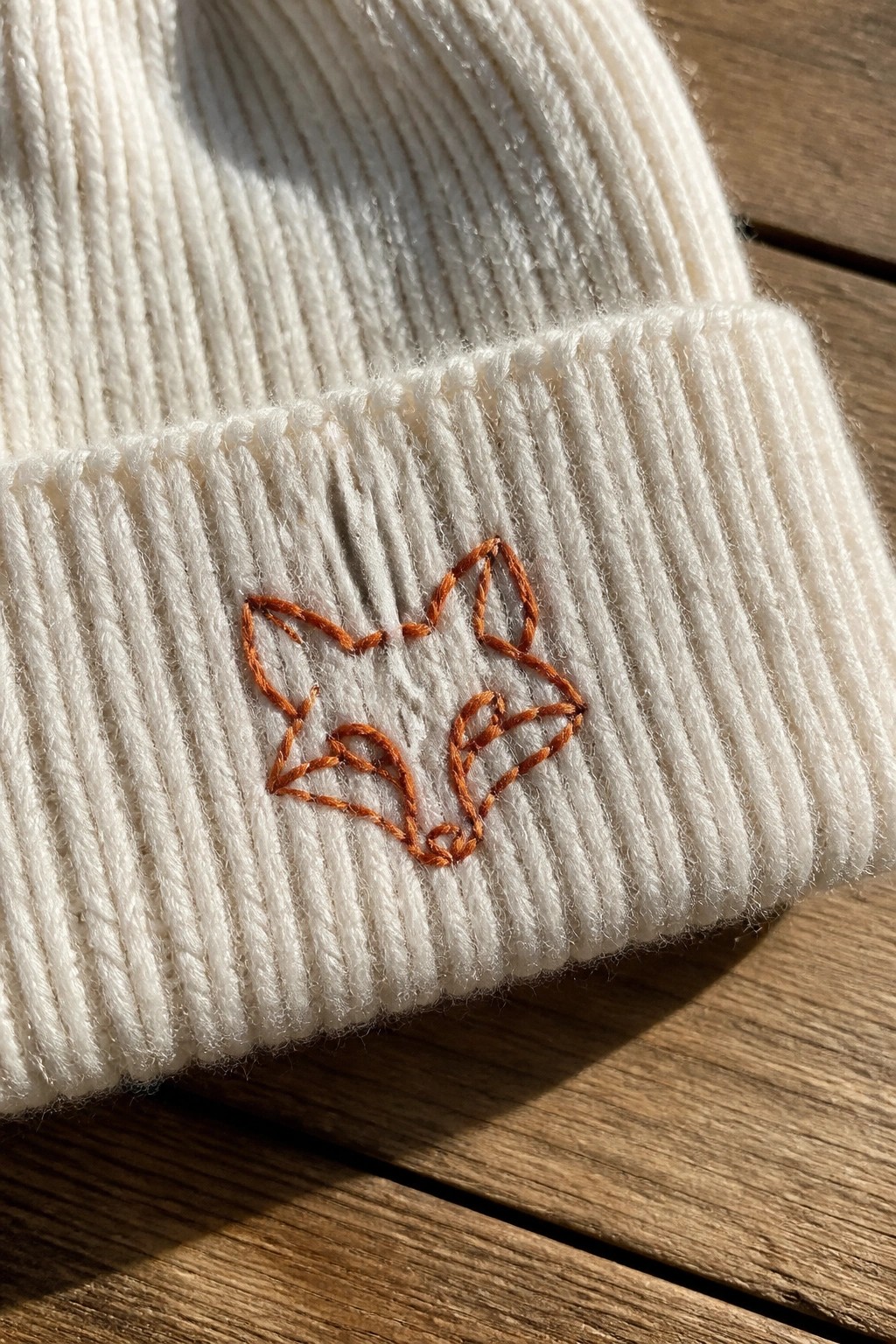 White ribbed beanie with orange embroidered fox on sunlit wood.