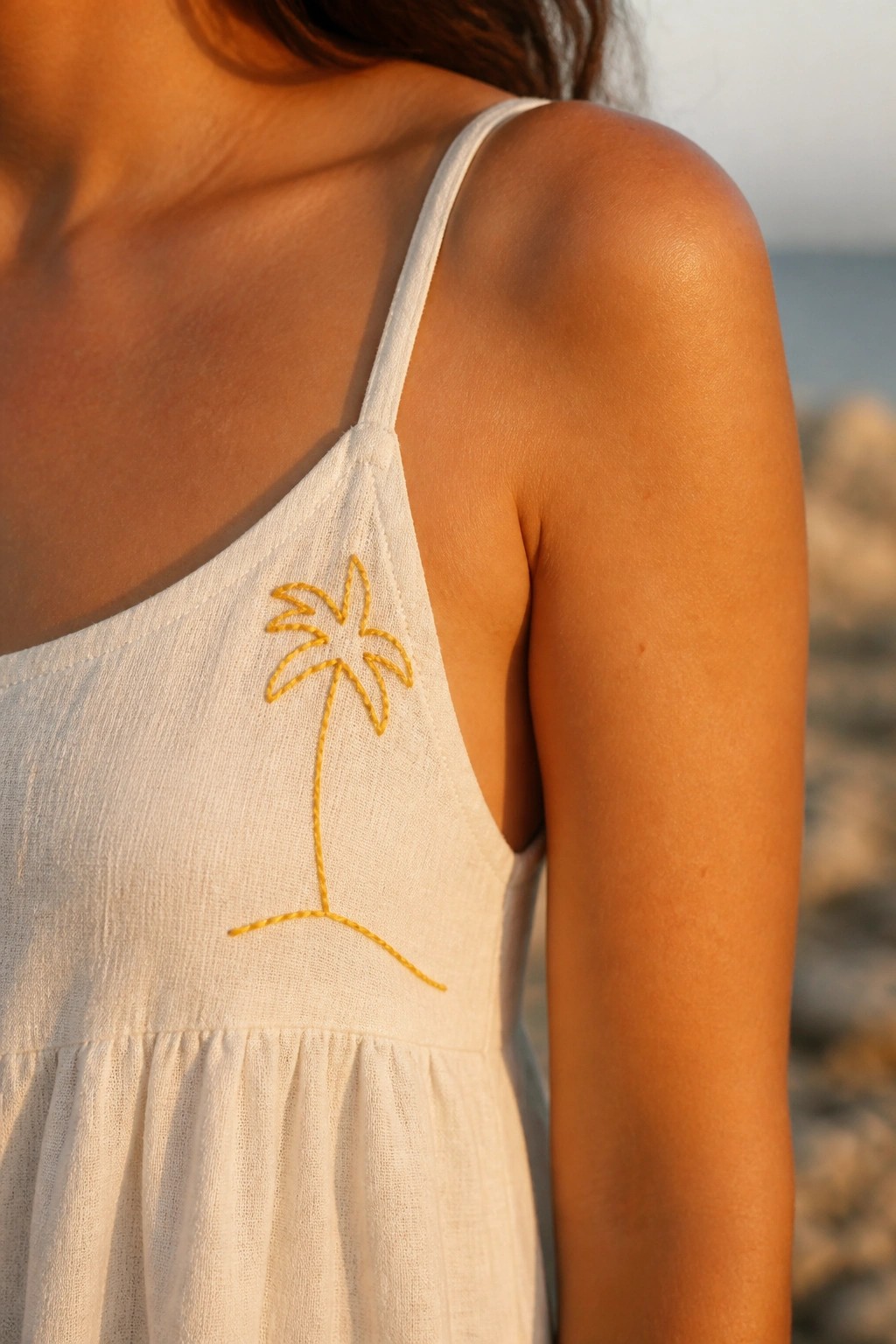 Close-up of a white linen top with a yellow single-line palm tree embroidered beside the spaghetti strap.