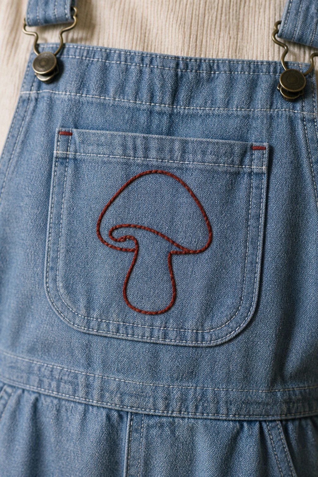 Reddish-brown mushroom outline embroidered on a denim overalls pocket.